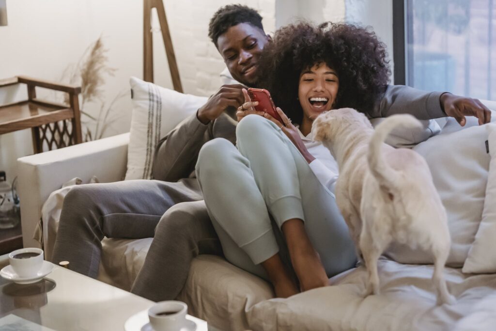 Delighted African American couple sharing smartphone on couch and playing with pet